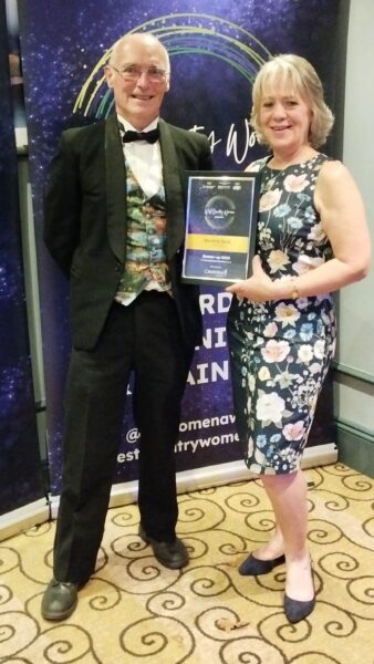 Michelle and Kevin in posh clothes standing in front of a West Country Women Awards Banner at the Awards Dinner